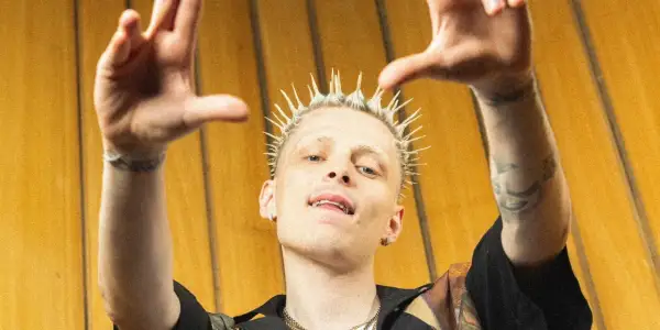 Raper Oki z blond irokezem, uniesionymi rękami i złotymi zębami na tle żółtej ściany.
