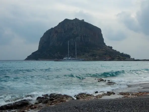 Monemvasia, Grecja: skalista góra z ruinami zamku na szczycie, jacht na turkusowym morzu i kamienista plaża.
