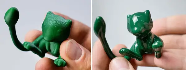 Due figurine verdi di gatto, stampate in 3D, tenute tra le dita. I colori per stampe 3d brillano sotto la luce.