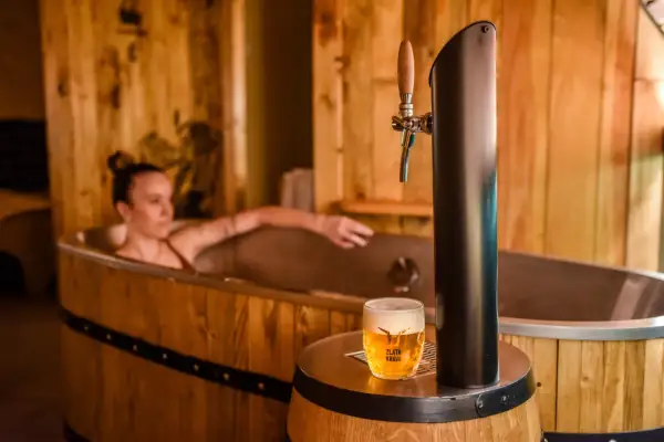 Entspannende Frau in einem Bierfass-Bad, daneben ein frisch gezapftes Pils. Ein einzigartiges Bier-Spa-Erlebnis in Pilsen.