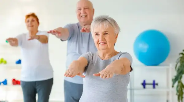 Trois seniors souriants pratiquent la gym chinoise pour seniors, bras tendus, dans une salle lumineuse.