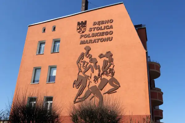 Pomarańczowa ściana budynku z płaskorzeźbą biegaczy i napisem "DĘBNO STOLICA POLSKIEGO MARATONU".