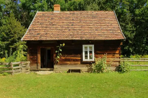 Drewniana chata z glinianym dachem, typowa dla skansenów w Polsce, otoczona zielenią i drewnianym płotem.