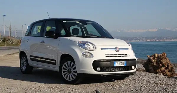 Biały fiat 500 l z czarnym dachem stoi na kamienistej plaży nad morzem.