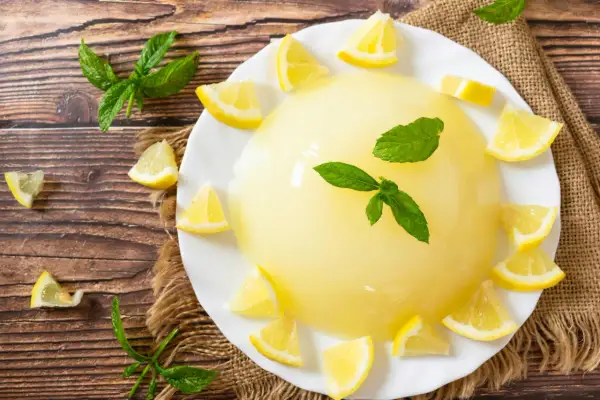 Un rinfrescante gelo di limone, guarnito con spicchi di limone e foglie di menta, su un piatto bianco, circondato da limoni e menta su un tavolo di legno.
