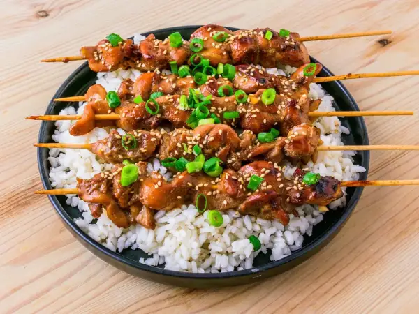 Fyra kycklingspett teriyaki ligger på en bädd av vitt ris, toppade med sesamfrön och hackad salladslök.
