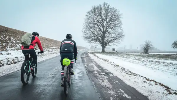 Dwaj rowerzyści na trasie Wisła 1200, w zimowej scenerii, pokonują oblodzoną drogę pod rozłożystym drzewem.