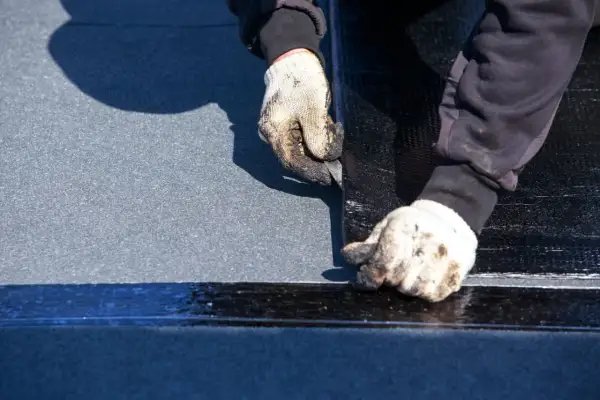 Manos enguantadas trabajando en la impermeabilización de tejados, cortando material negro sobre una superficie gris.