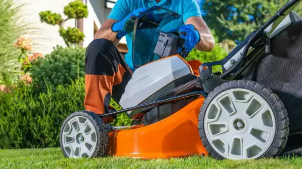 Mężczyzna wymienia baterię w pomarańczowej kosiarce akumulatorowej, przygotowując się do pracy w ogrodzie. To ważny element w rankingu kosiarek akumulatorowych.