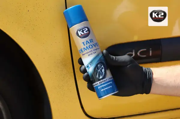 Rękawiczka z czarną gumą trzyma puszkę preparatu K2 Tar Remover, który skutecznie usuwa smołę z żółtego lakieru samochodu.