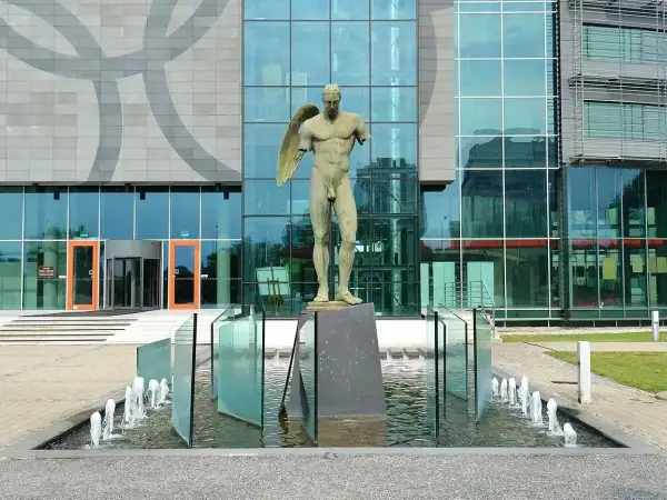 Brązowe rzeźby przedstawiające postacie ludzkie, w tym anioła, stoją przed nowoczesnym budynkiem z przeszkloną fasadą.