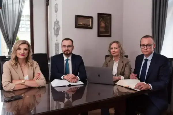 Cztery osoby w strojach biznesowych siedzą przy stole w sali konferencyjnej, przed nimi dokumenty i laptop.