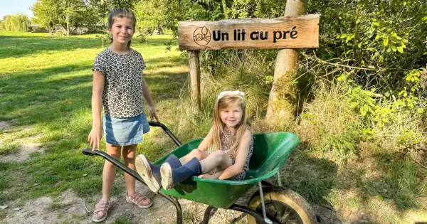 Deux petites filles profitent d'un séjour à la ferme, l'une assise dans une brouette, l'autre à côté, sous un panneau "Un lit au pré".