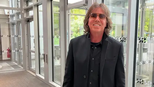 Joey Tempest, med sitt karaktäristiska hår och solglasögon, poserar framför en glasdörr.