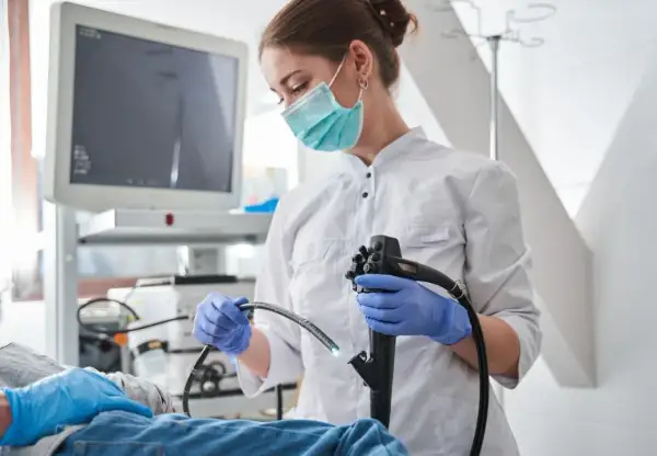 Lekarka w masce i rękawiczkach przygotowuje się do gastroskopii. Pacjent leży, a na ekranie widać obraz z kamery. Po zabiegu może pojawić się ból po gastroskopii.