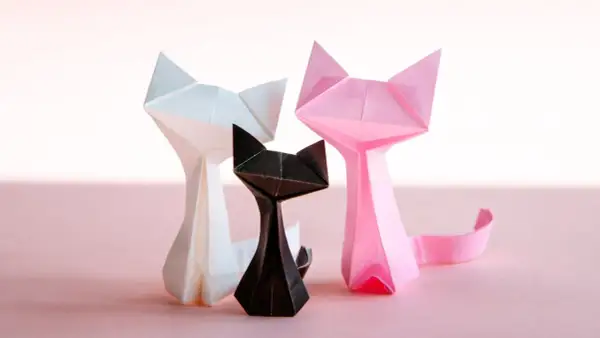 Trzy papierowe koty origami: biały, czarny i różowy, na jasnoróżowym tle.