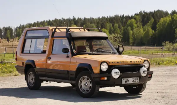En orange Talbot Matra Rancho står parkerad på en grusväg med skog i bakgrunden.