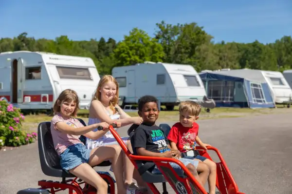 Fyra glada barn på en trampbil på Kolmården first camp. Husvagnar och träd i bakgrunden.