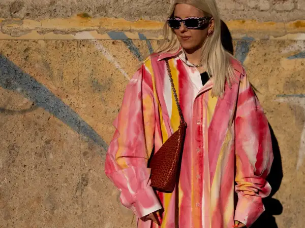 Kobieta w okularach przeciwsłonecznych i koszuli w stylu tie-dye w odcieniach różu i żółci, z brązową torebką na łańcuszku.
