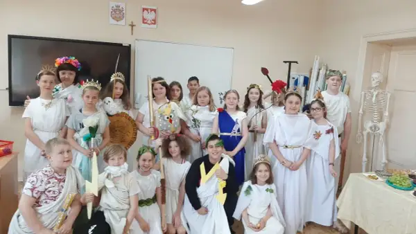 Dzieci w strojach antycznych bogów i bogiń, jak Artemida, pozują do zdjęcia. W tle szkielet i tablica.
