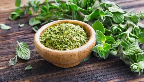 Un ciotola di legno piena di origano secco, con rametti freschi sullo sfondo. Il profumo di origano inebria l'aria.