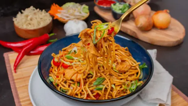 Bami Goreng mit Hühnchen, garniert mit Frühlingszwiebeln und Chili. Daneben sind Zutaten wie roter Chili, Knoblauch und Zwiebeln zu sehen.
