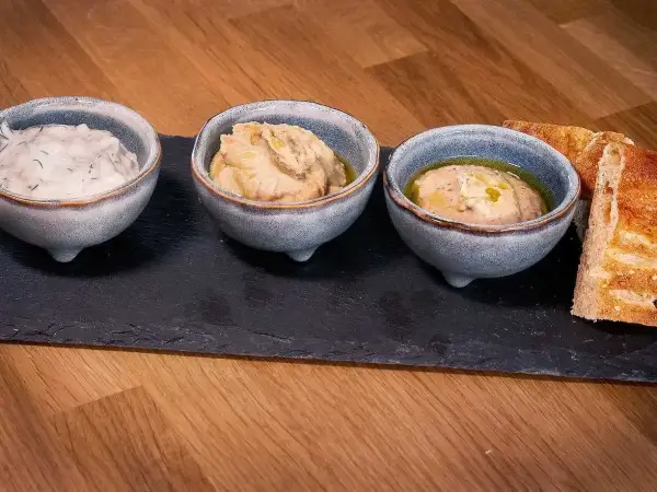 Tre skålar med dipp, en med dill, och två med en smak av 7 kryddor, serveras med rostat bröd på en stenplatta.