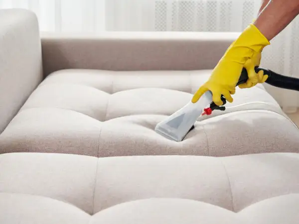 Gelbe Handschuhe führen ein Reinigungsgerät über ein helles Sofa. Tiefenreinigung für ein makelloses Sofa.