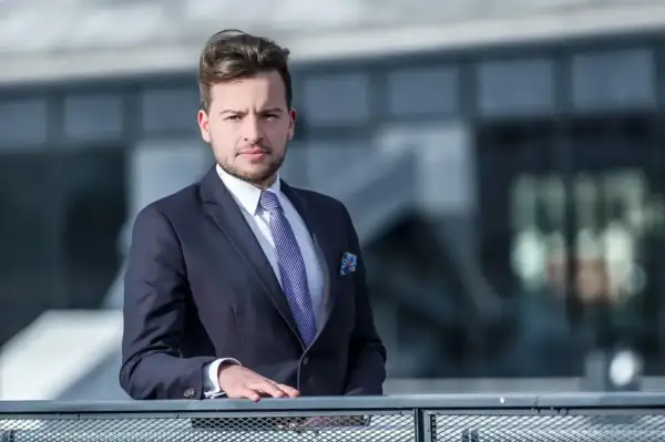 Mężczyzna w garniturze i krawacie opiera się o barierkę na tle nowoczesnego budynku.