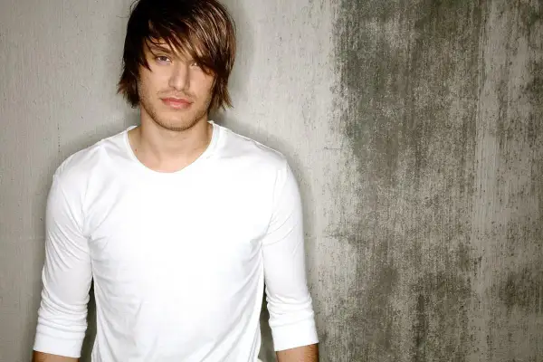 Michael Tyler, a young man with tousled brown hair and a white long-sleeved shirt, poses against a textured concrete wall.