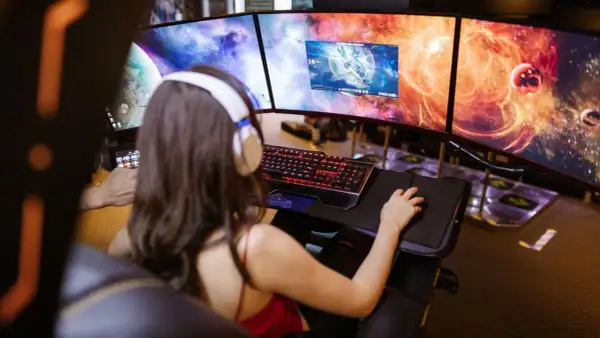 Frau mit Kopfhörern spielt an drei Monitoren. Ein "pile of shame" an Aufgaben wartet, doch jetzt zählt nur das Spiel.