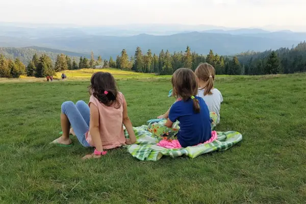 Trzy dziewczynki siedzą na kocu na trawie, podziwiając rozległy widok na Beskid Żywiecki.