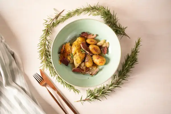Ein Teller mit Hähnchen, Kartoffeln und Speck, umrahmt von Rosmarinzweigen. Die Rosmarin verwendung verleiht dem Gericht ein wunderbares Aroma.