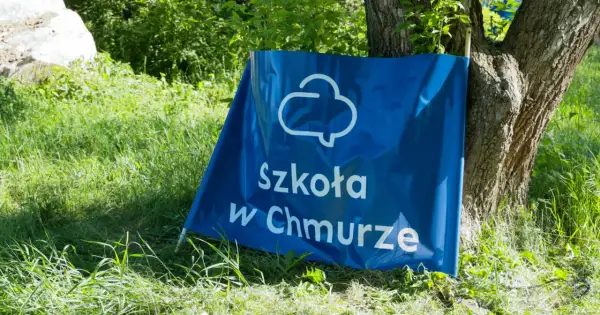 Niebieski baner z napisem "Szkoła w Chmurze" oparty o drzewo, na tle zielonej trawy i liści.