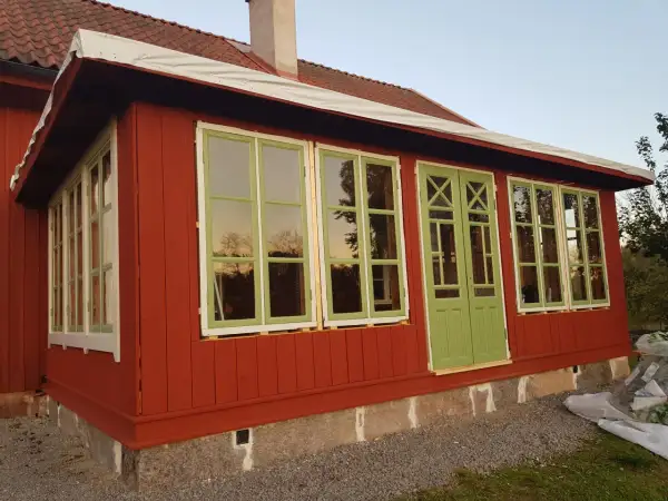 Ett rött hus med pågående arbete för att bygga glasveranda. Gröna fönster och dörrar reflekterar himlen.