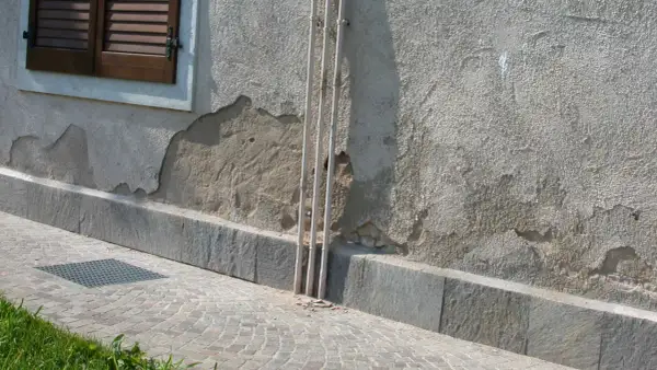 Il muro esterno grezzo mostra scrostamenti e necessita di essere pitturato. Tre tubi bianchi corrono verticalmente lungo la parete.