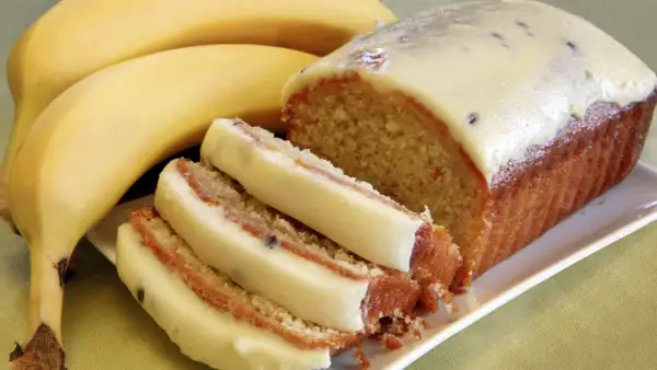 Wilgotne ciasto bananowe z kremową polewą i kawałkami owoców, obok dojrzałych bananów.