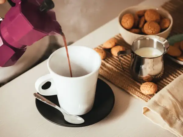 Un buon caffè fumante viene versato da una moka viola in una tazza bianca, accompagnato da biscotti e latte.