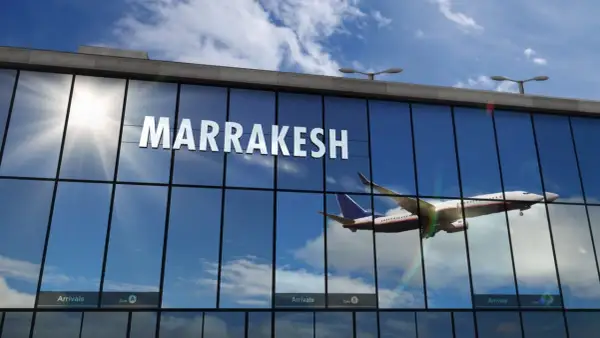 Samolot wznosi się na tle błękitnego nieba z chmurami, odbijając się w szklanej fasadzie lotniska w Marrakeszu.