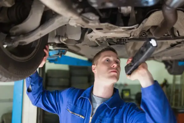 Mechanik w niebieskim kombinezonie bada podwozie auta latarką, przygotowując się do wymiany sworznia wahacza.