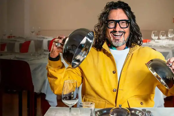 Chef con giacca gialla solleva un coperchio, pronto a svelare un piatto in un ristorante. Un assaggio di "4 ristoranti".