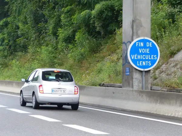 Voiture argentée sur l'autoroute, passant devant un panneau "Fin de voie véhicules lents". La nature luxuriante borde la route.