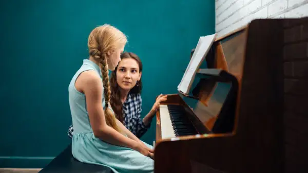 Dziewczynka z warkoczem uczy się gry na pianinie pod okiem nauczycielki. Nuty na pulpicie wskazują na lekcję muzyki.