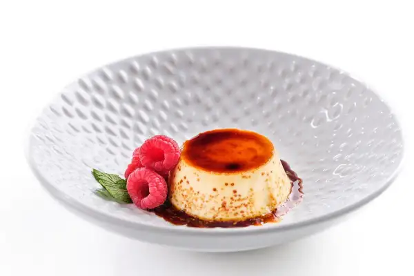 Delicioso flan de queso fresco con caramelo líquido, decorado con frambuesas y menta en un plato blanco texturizado.