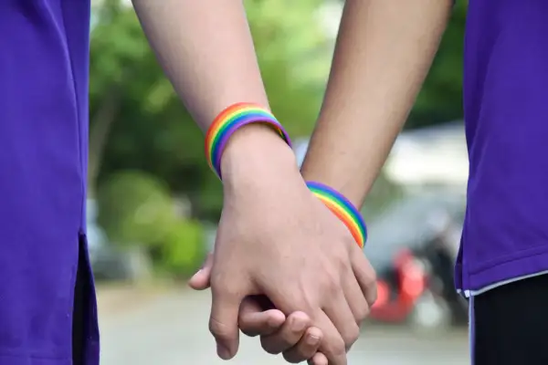 Zwei Hände halten sich, geschmückt mit Regenbogenarmbändern. Eine Person trägt ein lila Oberteil. Ein Symbol für Liebe, das halb Mann halb Frau sein kann.