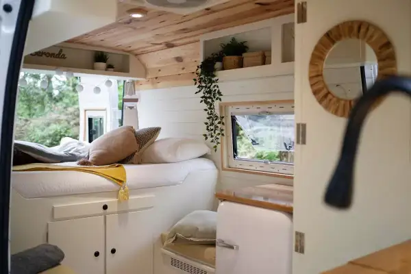 Interior de una camper con cama acogedora, cojines y mantas. Muestra cómo llevar la ropa de forma organizada en este espacio.