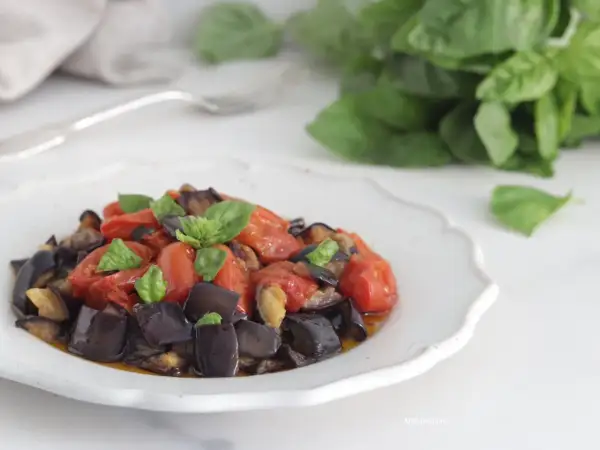 Melanzane a funghetto, un contorno saporito con pomodorini e basilico fresco, servito su un piatto bianco decorato.