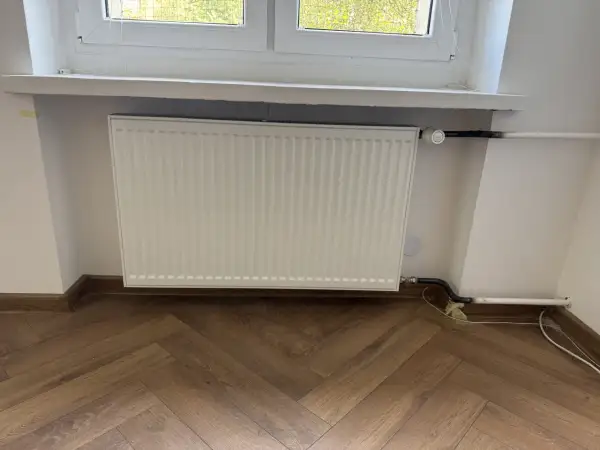 Biały grzejnik pod oknem, nowe podłączenie. Zastanawiasz się, ile kosztuje wymiana kaloryfera?