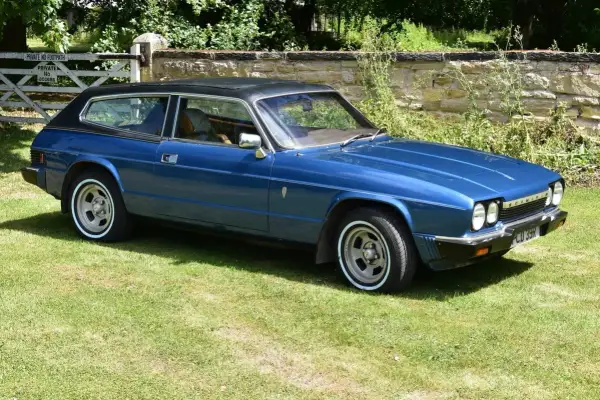 Blå Reliant Scimitar GTE, en klassisk sportsvagn med kombikaross, parkerad på en gräsmatta framför en stenmur och en grind.