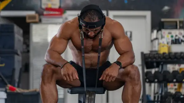 A muscular man sits, head harness on, preparing for neck exercises. He looks down, focused on his next move.